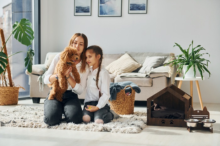 A mother and daughter play with a cute brown poodle in their living room.